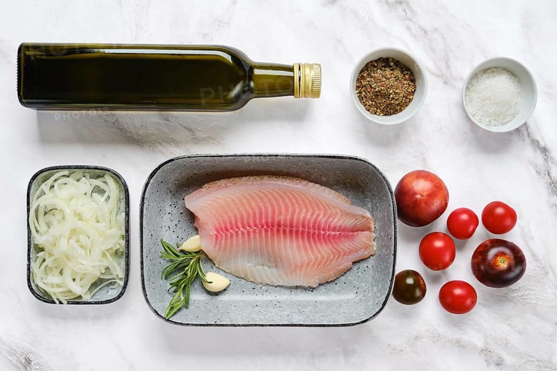 Fresh red seabream fish fillet on a gray plate surrounded by olive oil, tomatoes, onions, herbs, spices, and salt. Perfect setup for cooking a nutritious dinner in a modern kitchen