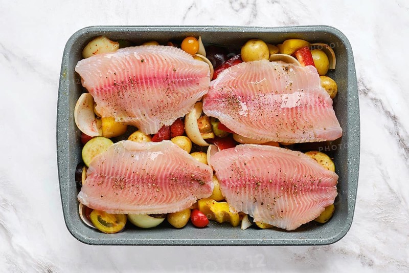 Fresh white fish fillets are arranged on a bed of colorful vegetables, including tomatoes and bell peppers. The dish is seasoned and prepared for a delicious baking in a roasting tray
