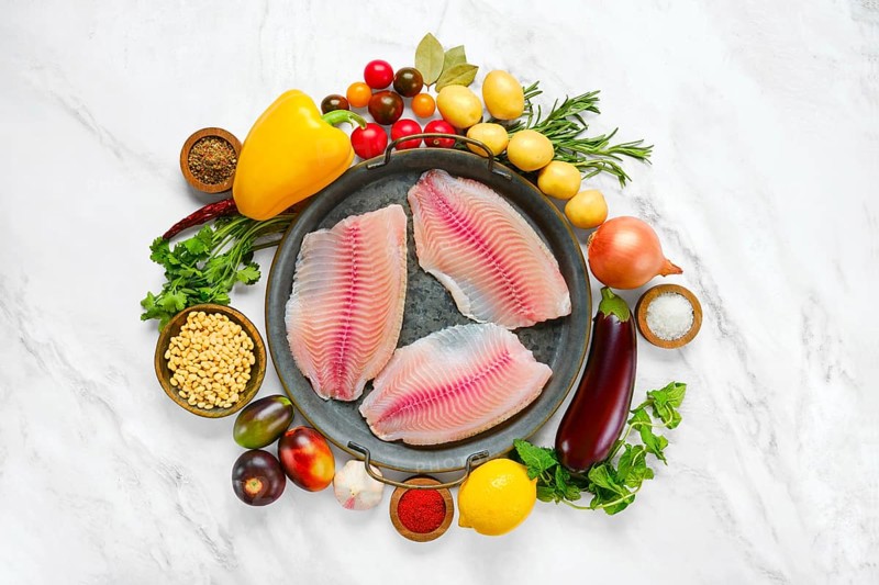 Three raw red seabream fish fillets on a round platter, surrounded by fresh vegetables like peppers, potatoes and tomatoes. Fresh herbs and spices add flavor, making it a perfect setup for cooking