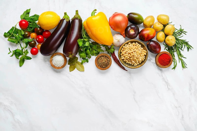 A colorful spread of fresh vegetables and herbs includes tomatoes, eggplants, cucumbers, bell peppers, potatoes, and various spices arranged on a light marble countertop in a kitchen