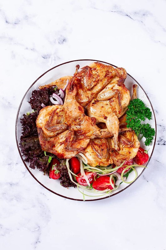 Baked quail, served on a plate with crispy lettuce leaves, fresh tomatoes and herbs, on a white plate, homemade