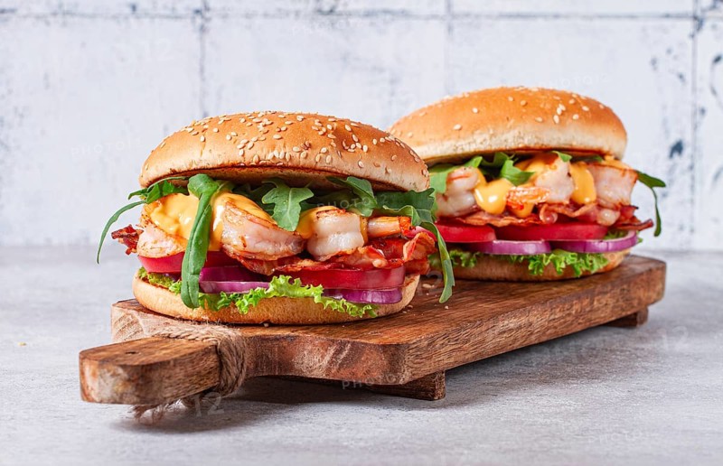 Two shrimp burgers and buns with sesame seeds, bacon, lettuce, tomatoes and onions, On a wooden board