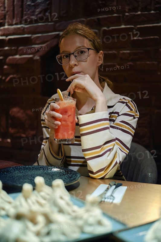 A young woman sits at a wooden table in a warm, dimly lit restaurant. She holds a colorful drink with a straw, gazing thoughtfully