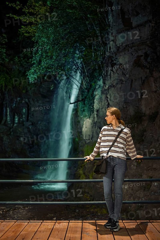 A young woman enjoys a quiet moment by a waterfall in a lush, natural setting during the evening hours. The scene is illuminated by soft evening light, enhancing the tranquil atmosphere