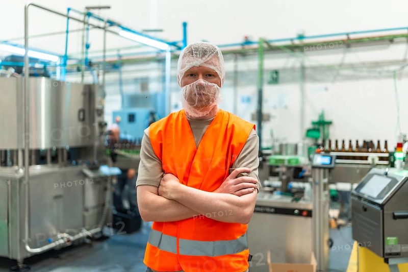 Worker standing proudly with crossed arms, wearing a safety vest and hairnet in a clean, automated beer bottling plant, overseeing the manufacturing process
