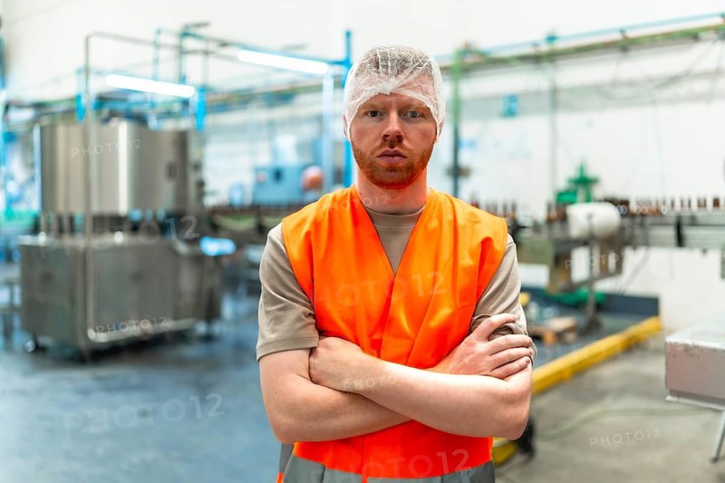 Male worker standing with crossed arms, wearing a hair net and a safety vest, overseeing operations in a food and beverage production facility with machinery in the background