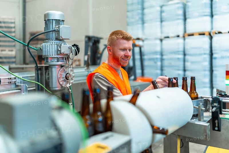 Smiling man in safety vest stands beside a conveyor belt, carefully inspecting brown glass bottles during the bottling process at a modern brewery factory