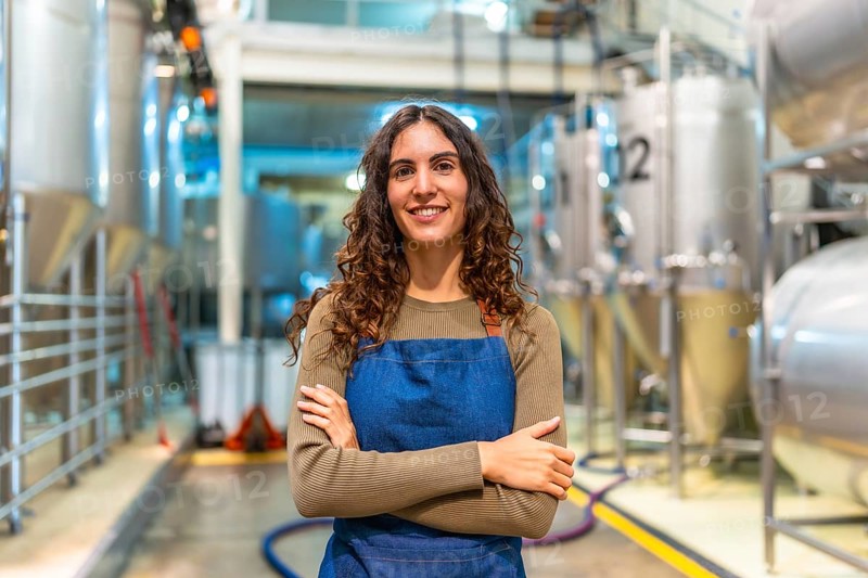 Woman brewer stands confidently with arms crossed, smiling at camera in a modern microbrewery with stainless steel fermentation tanks, proud small business owner and craftsperson