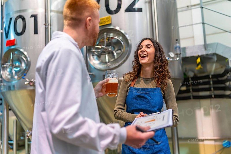 Brewery worker and quality control specialist smiling, holding clipboard and a glass of beer, discussing brewing process in a modern industrial facility