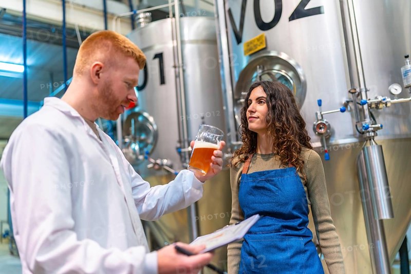 Brewery workers evaluating beer sample for quality control, holding clipboard and glass in brewing facility with large fermentation tanks, ensuring product excellence
