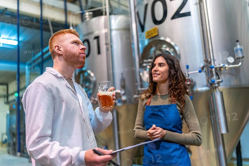 Two brewery professionals inspect and taste craft beer beside large fermentation tanks in a modern microbrewery, checking quality, recording results and collaborating on production