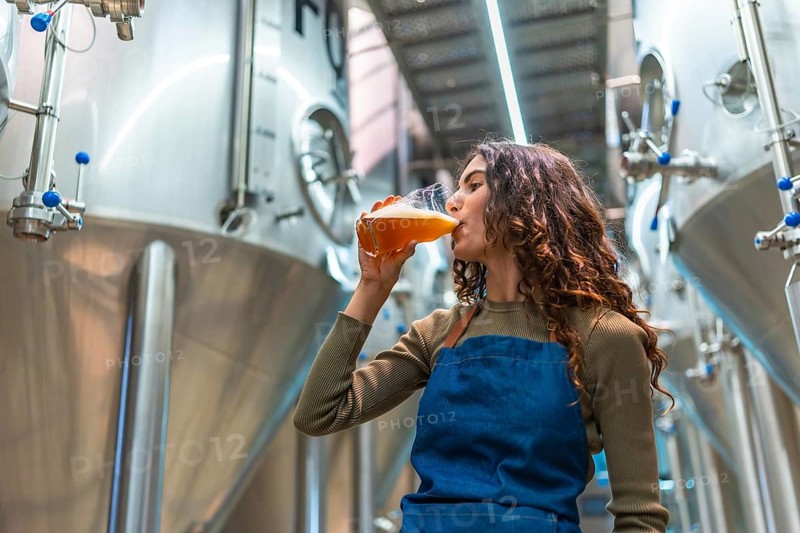 Woman wearing an apron tasting a glass of freshly brewed beer for quality control, standing in a modern micro brewery with large fermentation tanks in the background