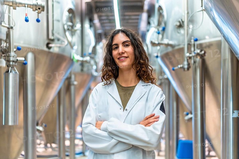 Woman wearing a lab coat standing confidently with crossed arms in a contemporary industrial brewery, surrounded by large stainless steel fermentation tanks
