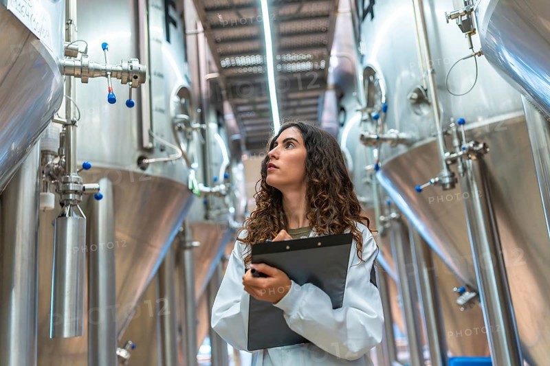 Woman in laboratory coat conducting quality control inspection in a modern beer brewery, holding a clipboard and looking up at rows of large stainless steel fermentation tanks