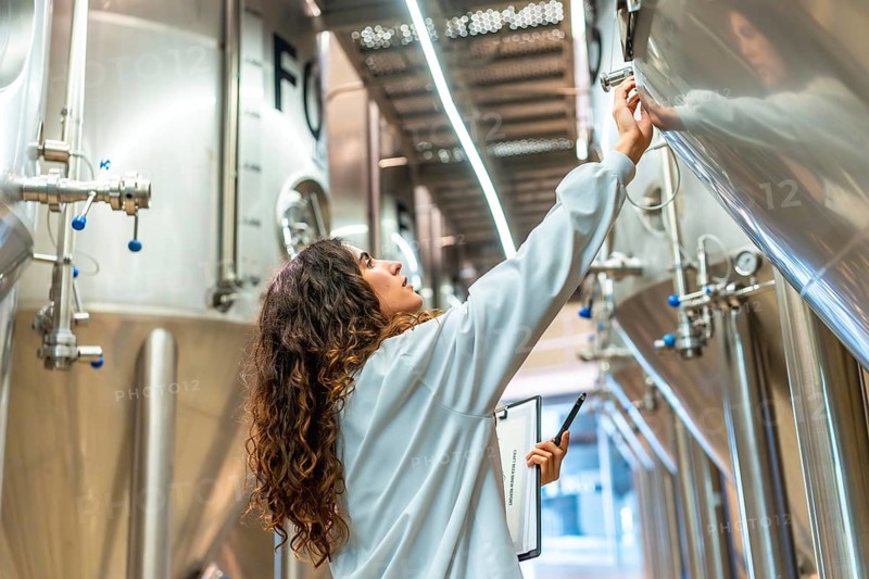 Young woman in a lab coat checking control panel on a stainless steel fermentation tank, performing quality control and inspection during the beer production process at a modern facility