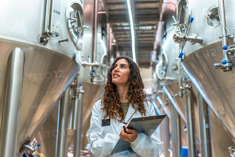 Woman scientist in a white lab coat with clipboard conducting quality control inspection in a modern craft beer production facility among stainless steel fermentation tanks and equipment