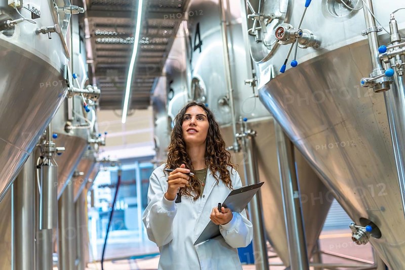 Woman wearing a lab coat holding a clipboard and pen, working in a modern brewery, inspecting stainless steel fermentation tanks and checking quality control