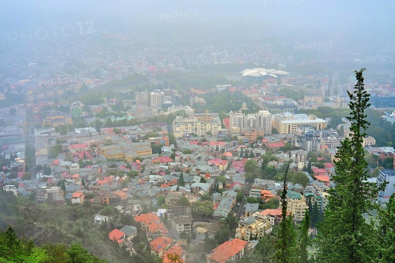 View of a city blanketed in fog with buildings and greenery in the foreground. Various buildings with colorful rooftops are visible, surrounded by trees and mountains