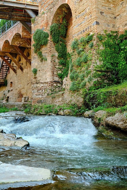 Clear water streaming over smooth stones next to an ancient stone wall adorned with lush plants. Sunlight illuminates the peaceful landscape, enhancing its natural beauty