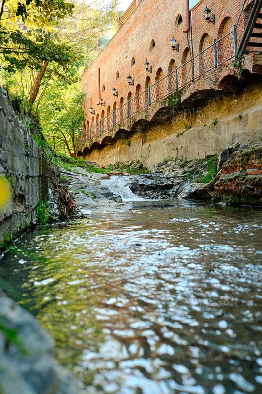 A clear stream flows calmly past old brick structures surrounded by lush greenery. Sunlight sparkles on the water's surface, creating a serene atmosphere in this peaceful outdoor scene