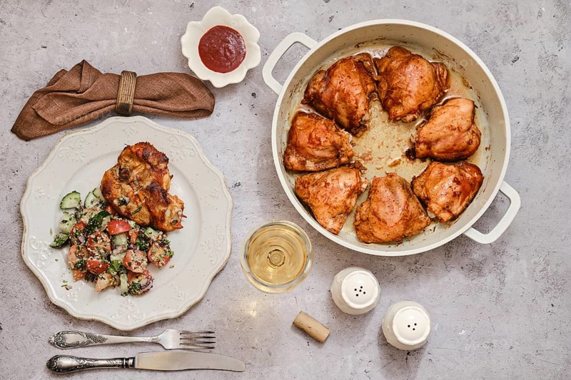 A well-cooked dish of roasted chicken thighs in a large pot, accompanied by a vibrant salad of cucumber and tomatoes on a plate, alongside a glass of white wine, ready for dinner