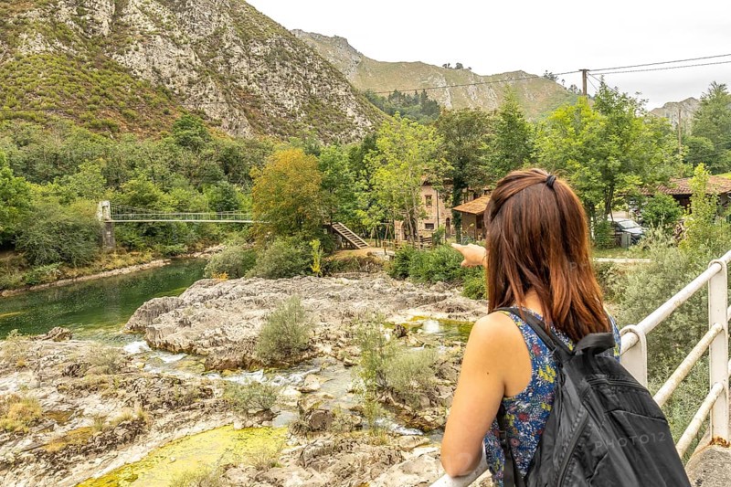 Tourist with a backpack pointing at the olla de san vicente, a natural pool on the sella river, surrounded by mountains and vegetation in sunny asturias, spain
