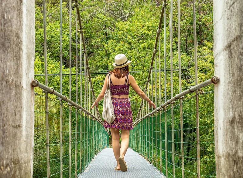 Young woman tourist with a hat and bag walking on a hanging bridge over the dobra river, enjoying a scenic summer adventure in asturias, spain