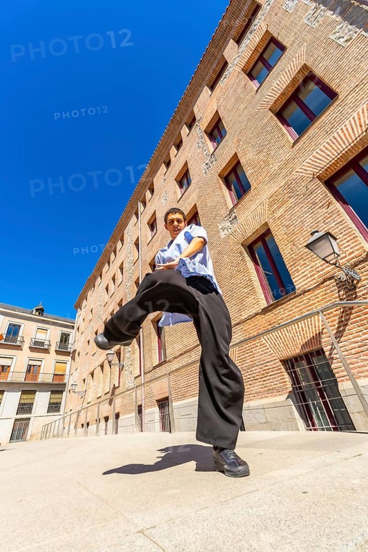 Young man doing a martial arts kick pose with focus and determination, dressed in casual loose clothing on a sunny day in an urban street with a brick building in the background