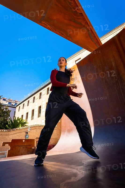 Young woman moving energetically, practicing dance in an urban setting, with modern corten steel architecture and a clear blue sky creating a dynamic scene