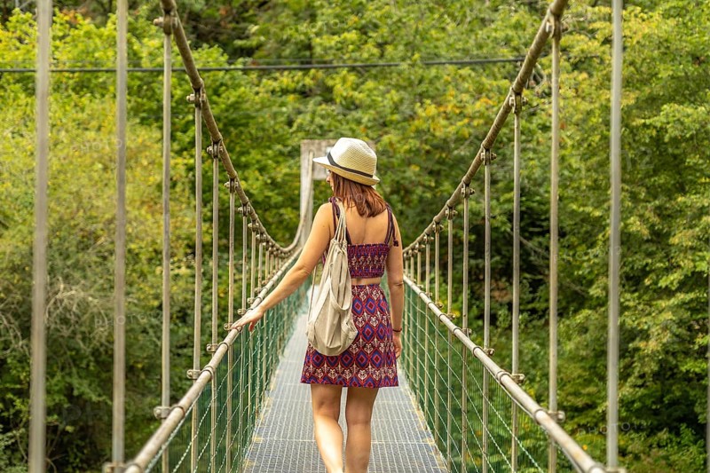 Female tourist with hat and backpack walking on a metal suspension bridge, enjoying the scenic beauty of a lush green forest, experiencing the thrill of adventure and exploration in nature
