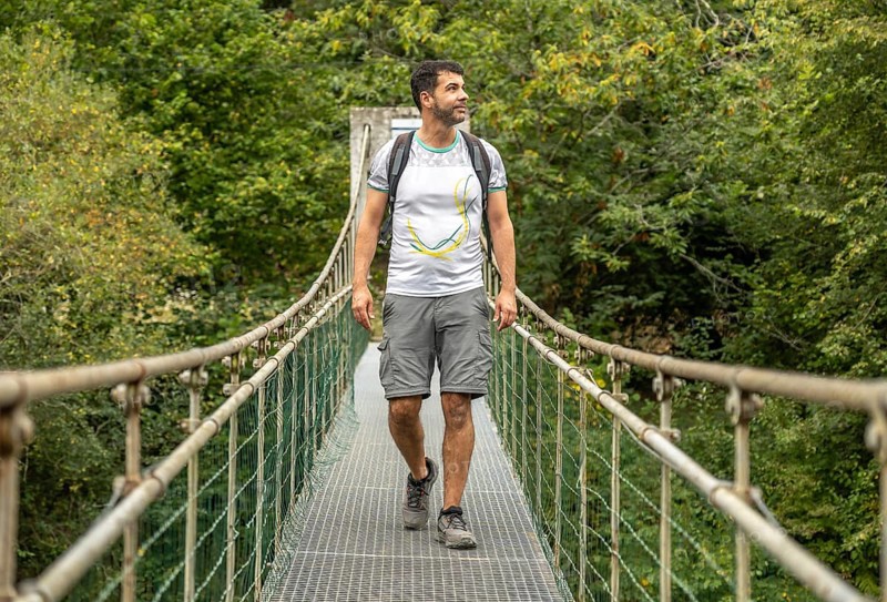 Male hiker with backpack enjoying trekking in the forest, walking on a metallic hanging bridge over a river, surrounded by lush green vegetation