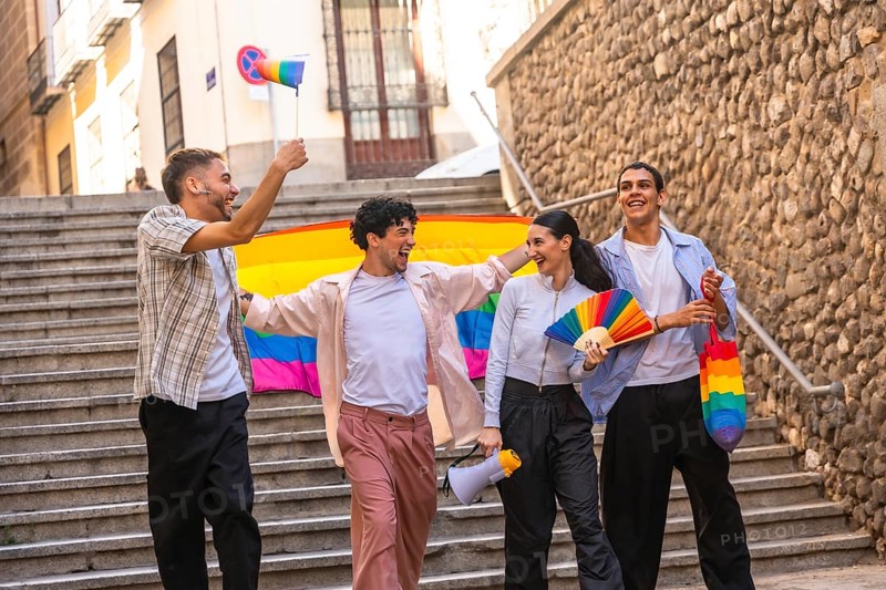 Diverse friendship group celebrating lgbtq plus pride and equality during a parade, walking down urban stairs, holding a rainbow flag, fan, megaphone, and small flag, showing joy and community
