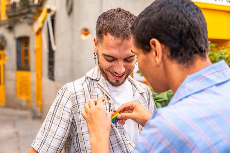 Young gay man gently attaching a rainbow pride flag pin to the plaid shirt of his smiling partner, celebrating love and lgbtq plus rights on a city street