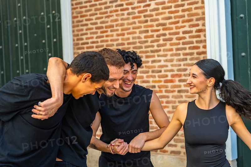 Diverse group of young adults laughing and embracing outdoors, enjoying casual summertime hangout and genuine friendship against an urban brick wall backdrop, carefree and joyful