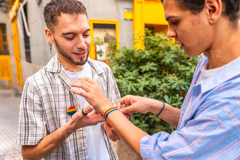 Two men clasp hands and put on rainbow flag bracelets outdoors, celebrating love, pride and identity with affectionate, candid street style connection and joyful solidarity