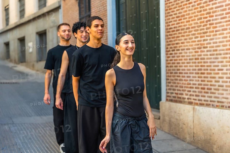 Young diverse dancers walking in line down a city street, smiling and energized while rehearsing choreography outdoors against brick buildings, joyful teamwork and movement