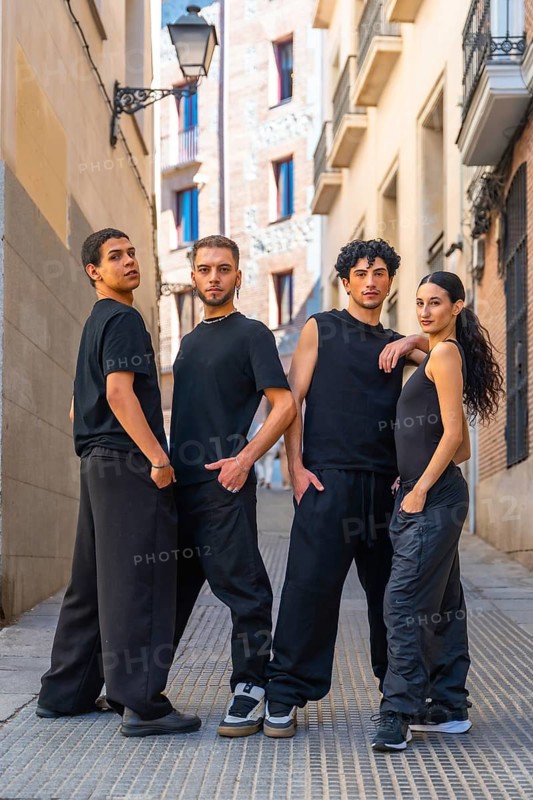 Four young adults in coordinated black streetwear pose confidently on a cobblestone alley, full length group portrait blending urban fashion, dance attitude and close knit friendship