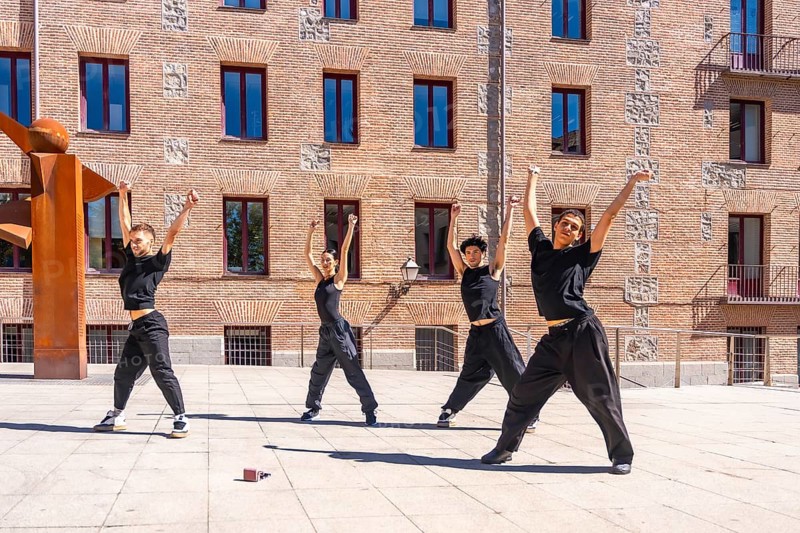 Four young male and female dancers performing a synchronized urban choreography with arms raised, expressing passion and energy against a brick building backdrop on a sunny day