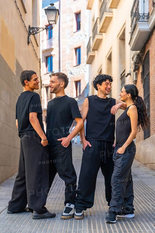 Four diverse young friends wearing black casual clothing are happily interacting, smiling, and talking while standing together in an urban alley or street