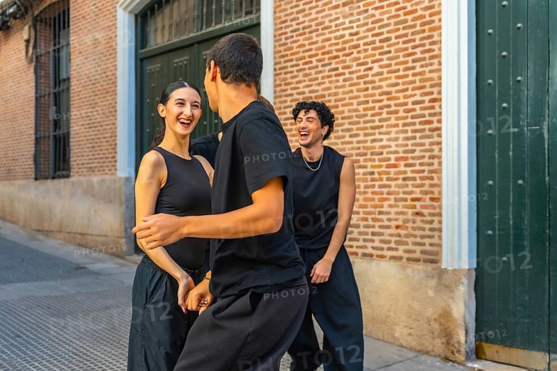 Young friends in black outfits laughing and dancing together on a city street by a brick wall, sharing joy, energy and playful urban togetherness in daylight