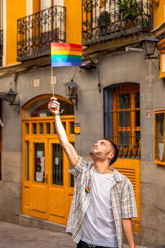 Young man standing in a city street, raising a small rainbow flag with arm outstretched, celebrating lgbtq plus pride, freedom, community and joyful support during daytime urban scene