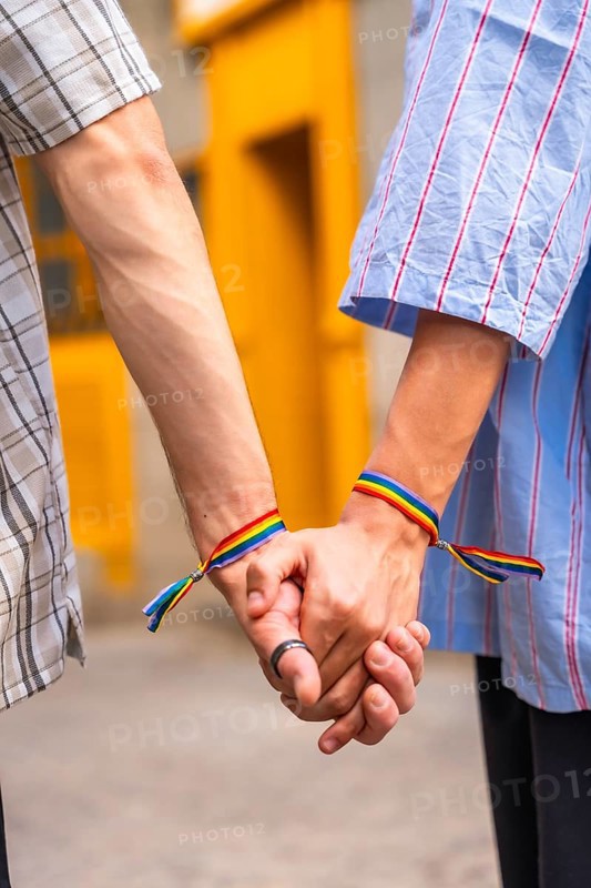 Male couple showing love and support for each other by holding hands with rainbow flag bracelets, symbolizing their lgbtq plus pride and relationship against a vibrant yellow background