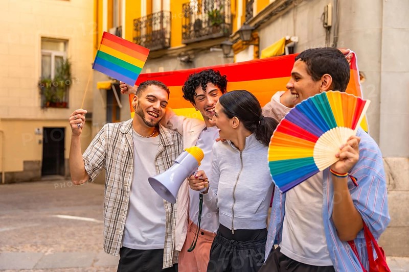Group of diverse young adults laughing and holding a rainbow flag, a small rainbow flag, a rainbow fan, and a megaphone, expressing joy and unity during a pride celebration