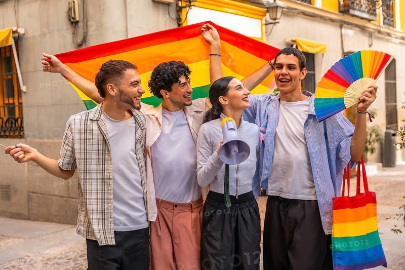 Diverse group of young adult friends smiling and holding a rainbow flag, fan, megaphone, and tote bag, celebrating pride and their community in an urban setting