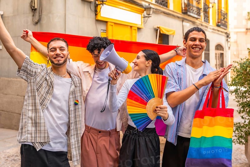 Diverse group of young friends standing together, holding a rainbow flag, fan, and megaphone, joyfully celebrating lgbt pride, diversity, and equality on a city street