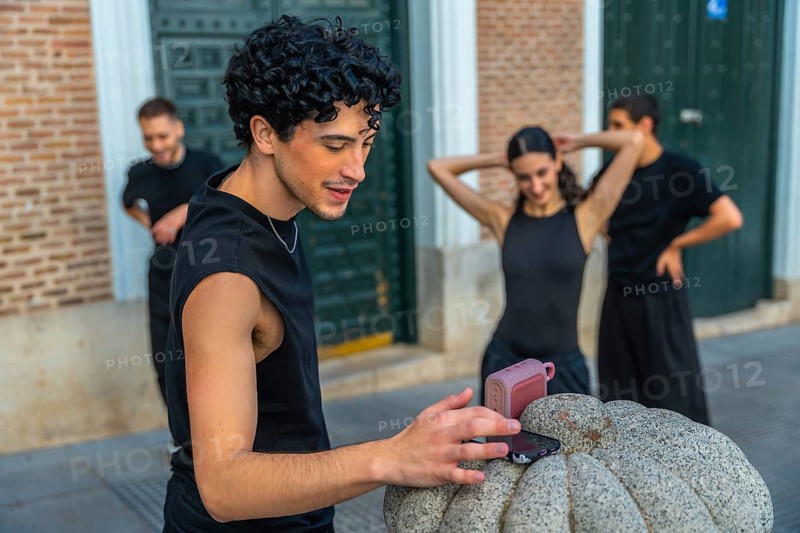 Young man playing music from a smartphone and portable speaker as urban dancers warm up on a city street, sharing rhythm, friendship and modern street culture