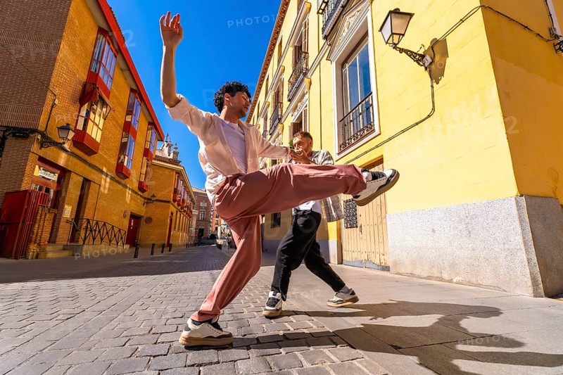 Two energetic young men are performing a dynamic dance routine on a cobblestone street, expressing happiness and freedom as bright sunlight illuminates the colorful urban architecture