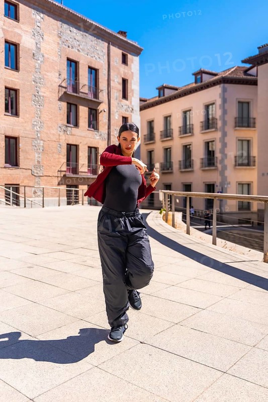 Young woman dancer performing dynamic contemporary routine on a sunny city street, showcasing urban fashion, confident expression and athletic movement against architecture