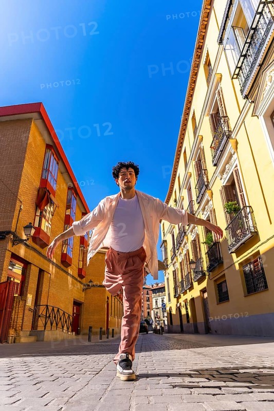 Young man actively dancing with outstretched arms on a sunny cobblestone street, expressing joy and freedom in a lively city environment with traditional buildings