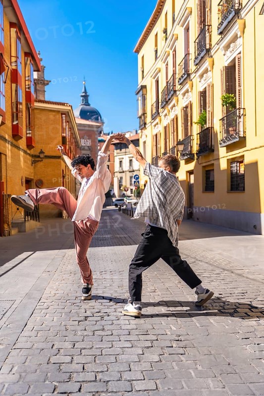 Two energetic young men performing a street dance on a cobblestone street in a lively european urban setting under clear blue skies, expressing creativity and freedom
